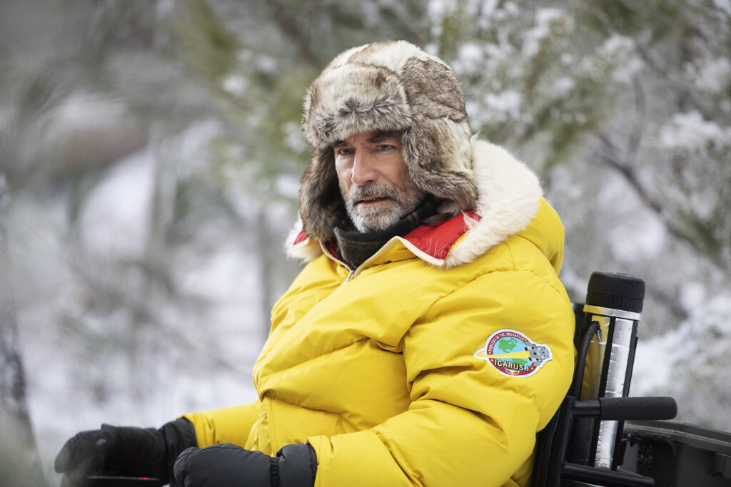 Niles Caulder dressed for intense cold in his wheelchair in a snowy forest.