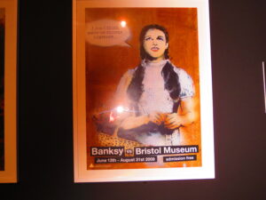 Banksy vs Bristol Museum poster of a spray-painted Dorothy from the Wizard of Oz saying “I don’t think we’re on canvas anymore…”
