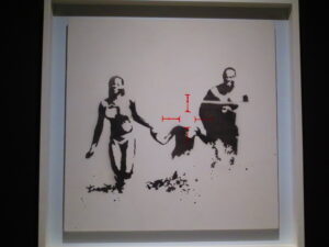 Family Target by Banksy. Spray painted on board, a happy family holding hands, with a bomb target where the child’s face is