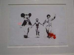 Napalm, Serpentine Edition by Banksy. Silkscreen print of an altered version of “The Terror of War (Napalm Girl)” photograph by Nick Ut. The titular girl is naked, screaming from burns, and running, this time holding hands with Mickey Mouse and Ronald McDonald. There is a bloodstain smeared on the legs of Ronald.