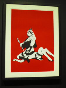 Queen Vic Screenprint by Banksy. On red background Queen Victoria is holding a staff and wearing a crown while sitting on top of a woman’s head who is in lingerie and heels