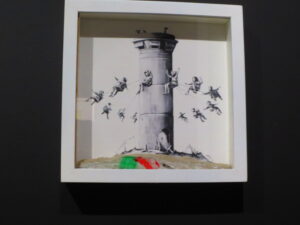 Walled Off Hotel Box Set by Banksy. Digital print on paper and wall section features children having fun on a carousel but it’s a watchtower resembling those found along the entire separation wall