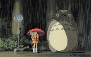 two girls under an umbrella standing beside Totoro, a large fantasy creature with a round belly and pointy ears