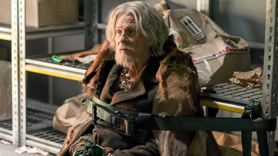 A shaggy and grey haired Silas has a green harness wrapped around him that's faceted to a metal shelf, completely immobilizing him.