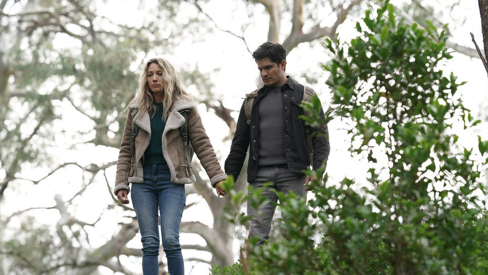 Eve and Levi facing the audience as they hold hands in a forrest as Eve looks down at something. Shrubbery to the right, partially covering Levi.