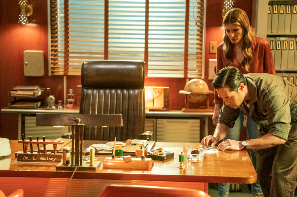 Ben as the janitor and Addison inspect a piece of paper in a 1960s office