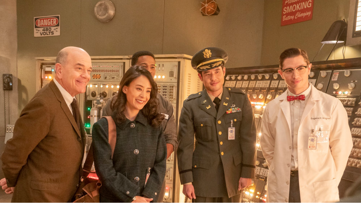 Dr. Woolsey, Mallory, Moe, Eugene, and Ben as the Colonel stand and smile in a nuclear reactor control room