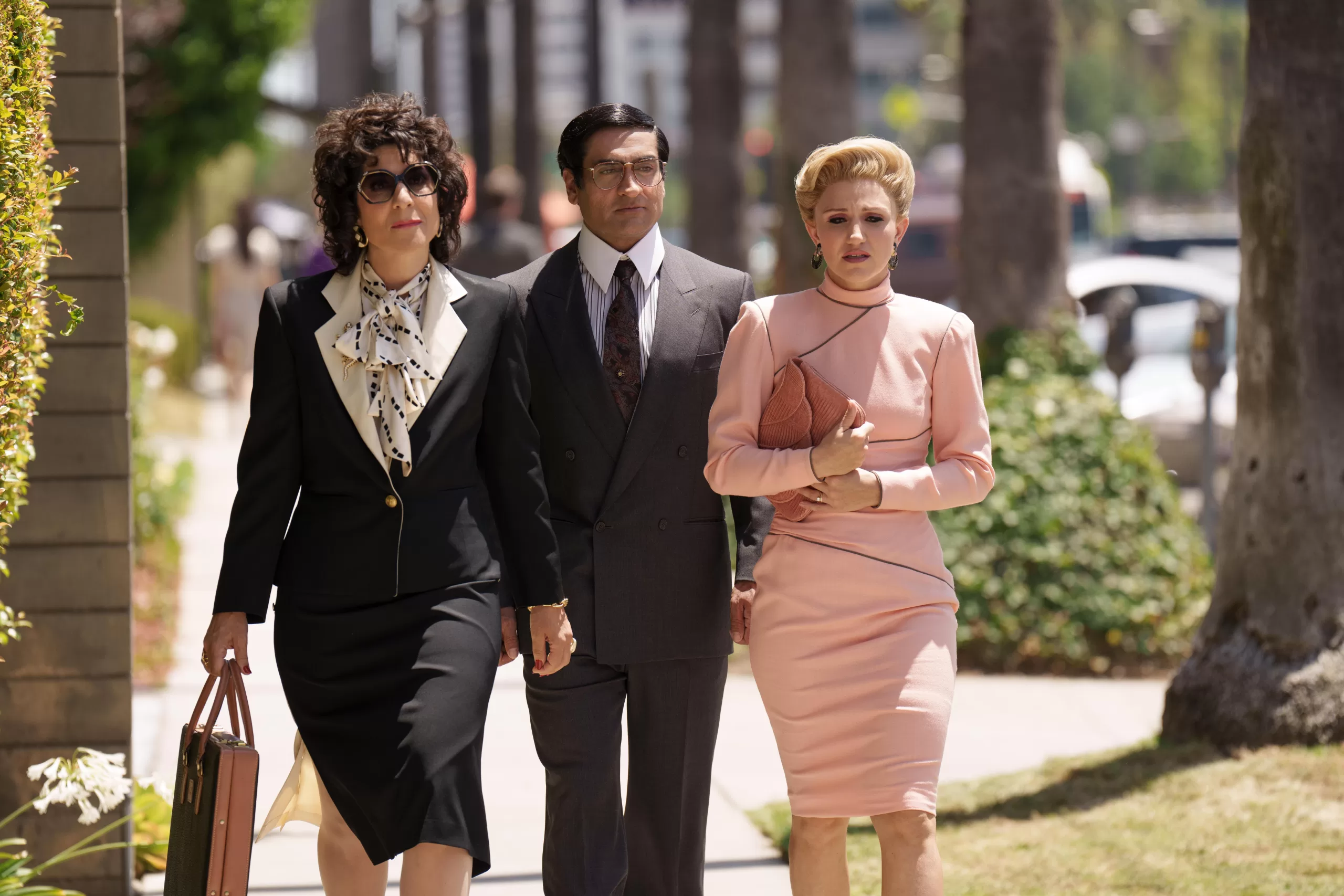 Kumail in a suit as Steve walks towards the camera, with Cheryl to his left in a black suit, and Irene to his right in a pink suit. All of them are dressed in 80s attire.